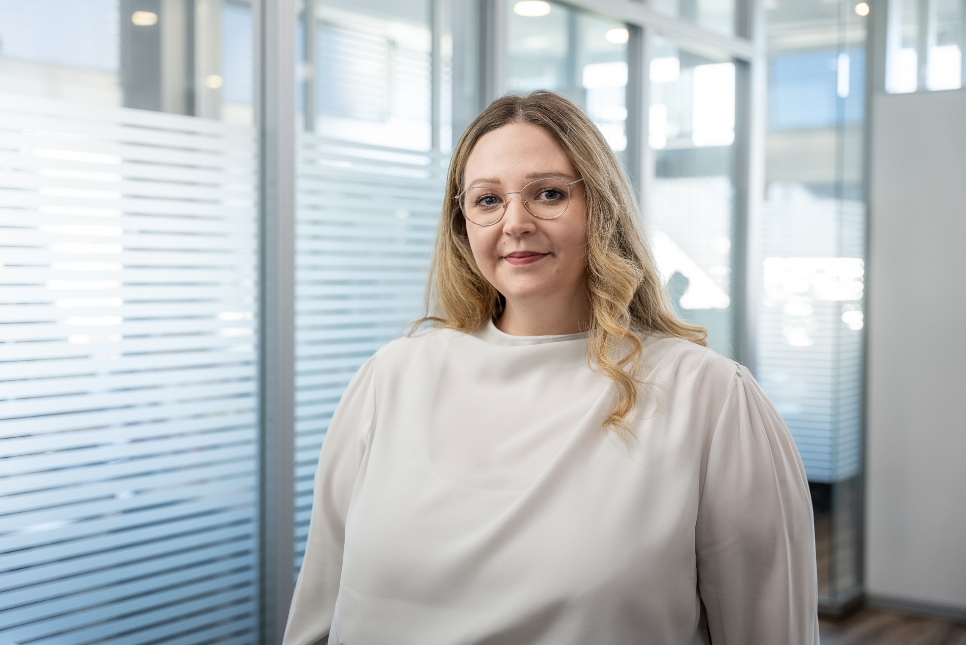 Eine Person mit langen, blonden, lockigen Haaren und einer hellen Bluse steht in einem modernen Büroumfeld mit Glaswänden und natürlichem Licht.