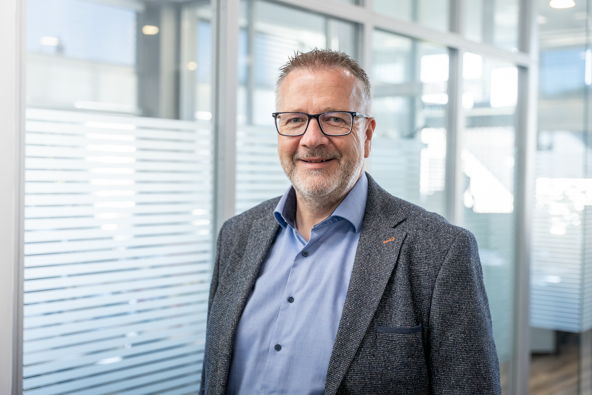 Ein Mann mittleren Alters mit Brille und grauem Bart steht in einem modernen Büroumfeld mit Glaswänden und verschiedenen Lichtquellen. Er trägt einen grauen Blazer und ein hellblaues Hemd, lächelt und sieht freundlich aus.