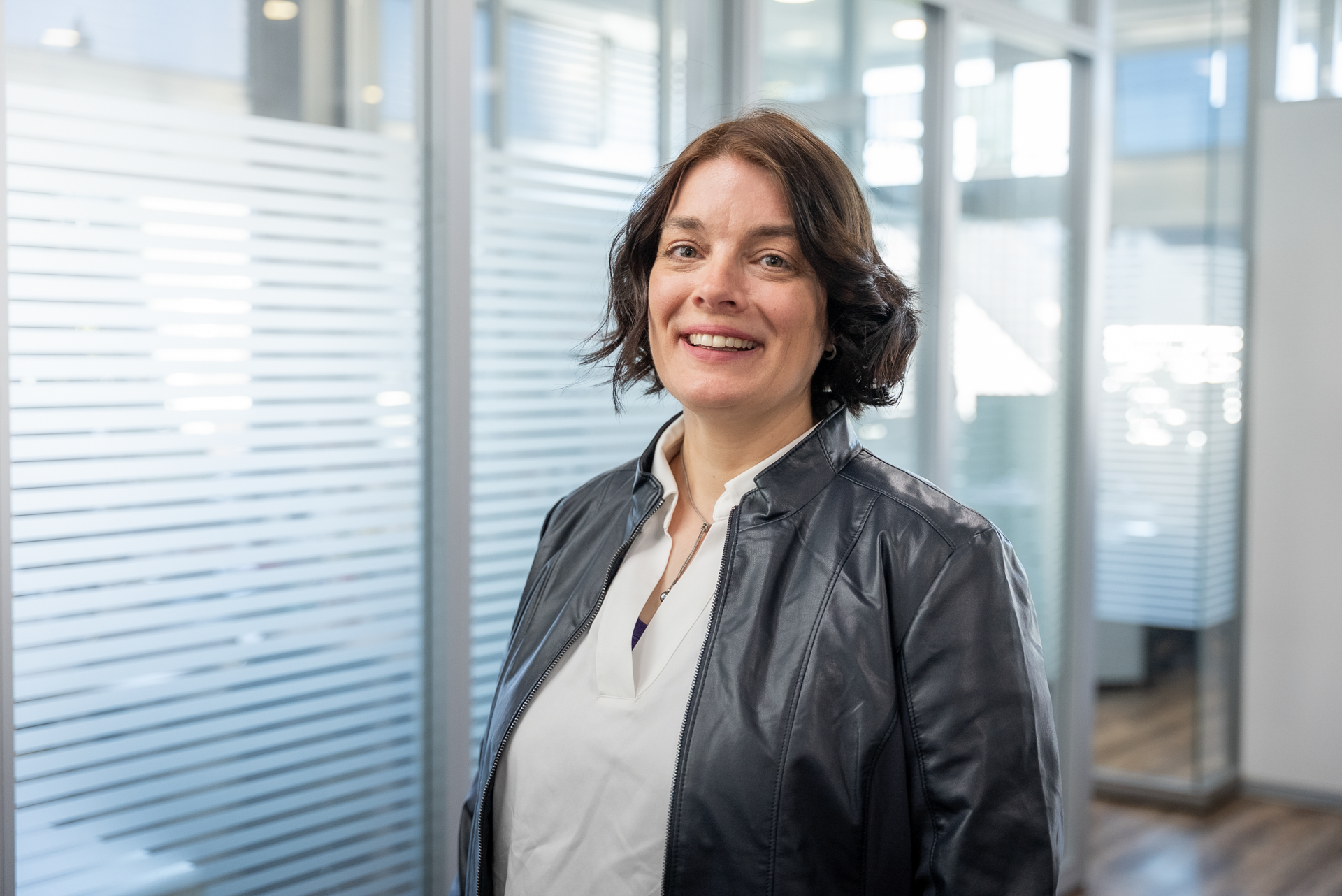 Eine lächelnde Frau mit dunklen, lockigen Haaren trägt eine schwarze Lederjacke und steht in einem modernen Büro mit Glaswänden.