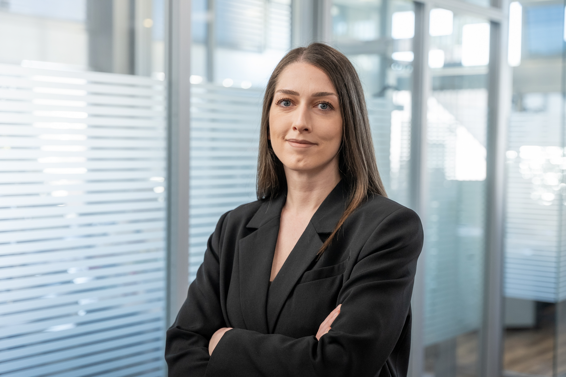 Eine Frau mit langen, glatten Haaren steht in einem modernen Büro. Sie trägt einen schwarzen Blazer und hat die Arme vor der Brust verschränkt, während sie freundlich in die Kamera schaut. Im Hintergrund sind Glaswände und helles Licht zu sehen.