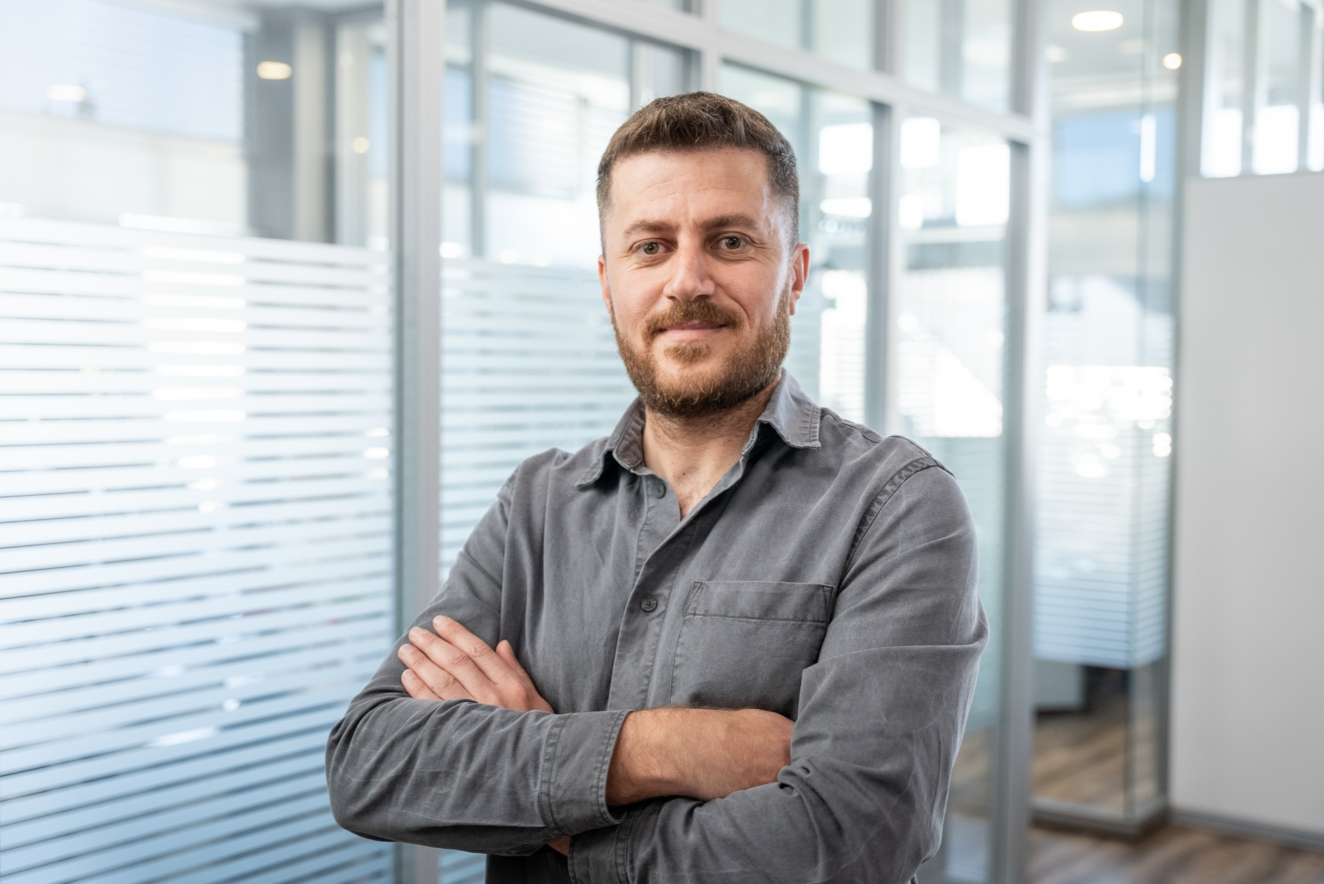 Ein Mann mit Bart in einem grauen Hemd steht mit verschränkten Armen in einem modernen Büro mit Glaswänden.