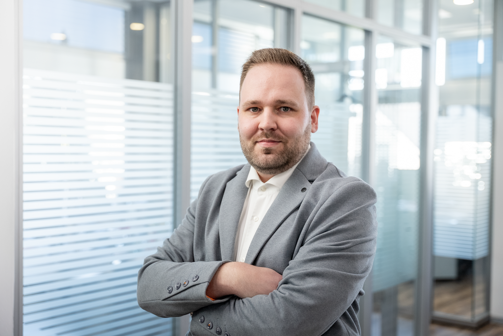 Ein Mann mit kurzen Haaren und einem grauen Sakko steht verschränkt in einem modernen Büro mit Glaswänden.