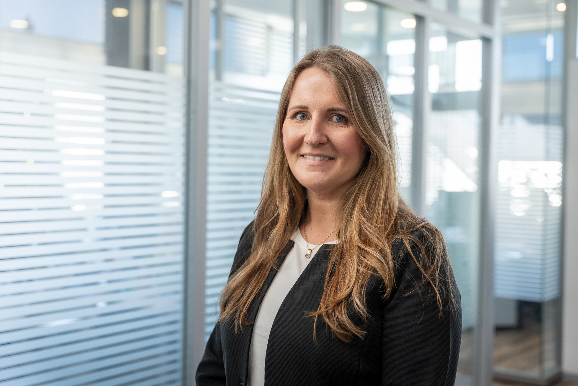 Eine lächelnde Frau mit langen, blonden Haaren trägt einen schwarzen Blazer und steht in einem modernen Büroraum mit Glaswänden und einem hellen, freundlichen Ambiente.
