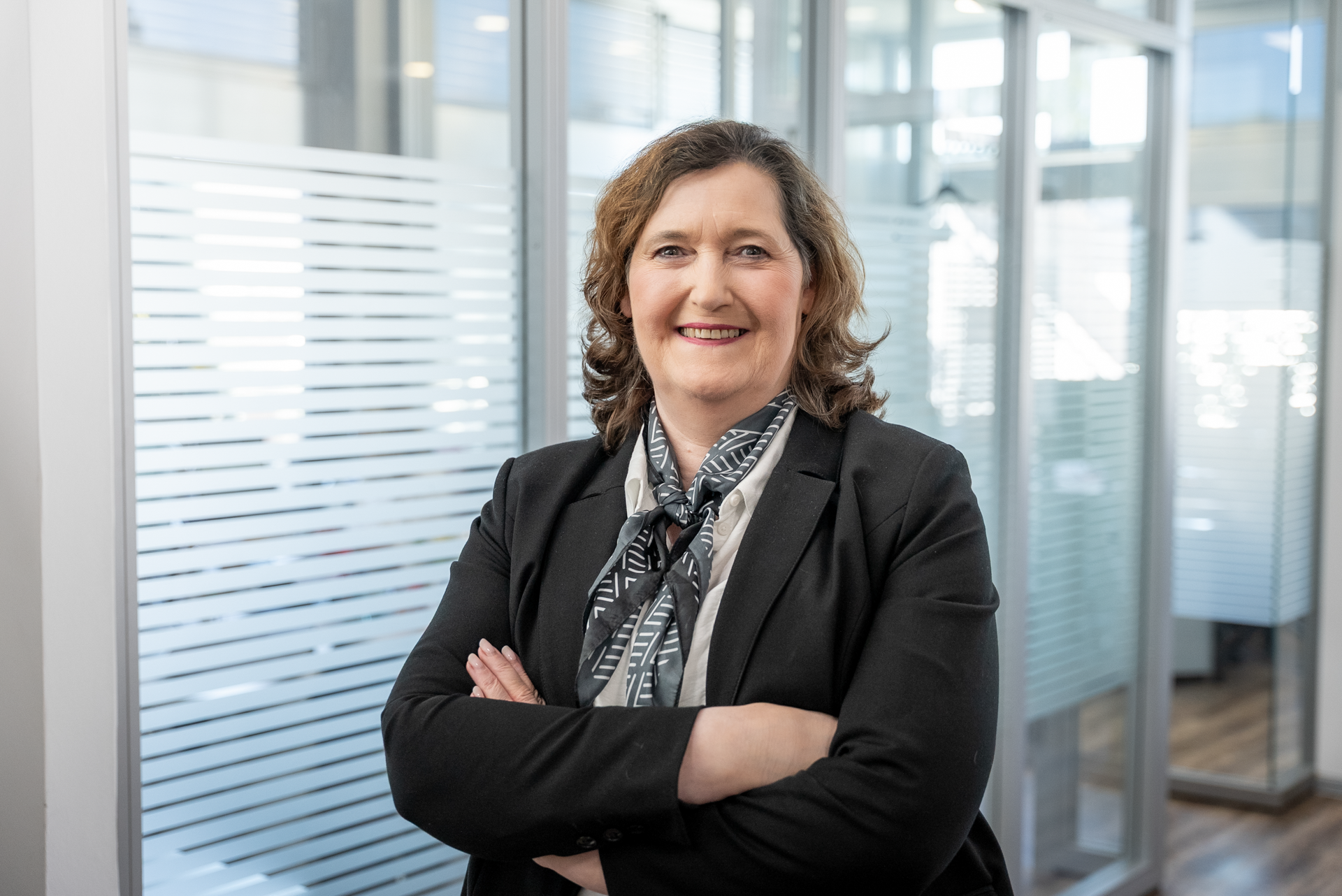 Eine beruflich gekleidete Frau mit lockigem, braunem Haar lächelt, während sie in einem modernen Büroraum mit großen Fenstern und Glaswänden steht. Sie hat die Arme verschränkt und trägt ein gestreiftes Halstuch.