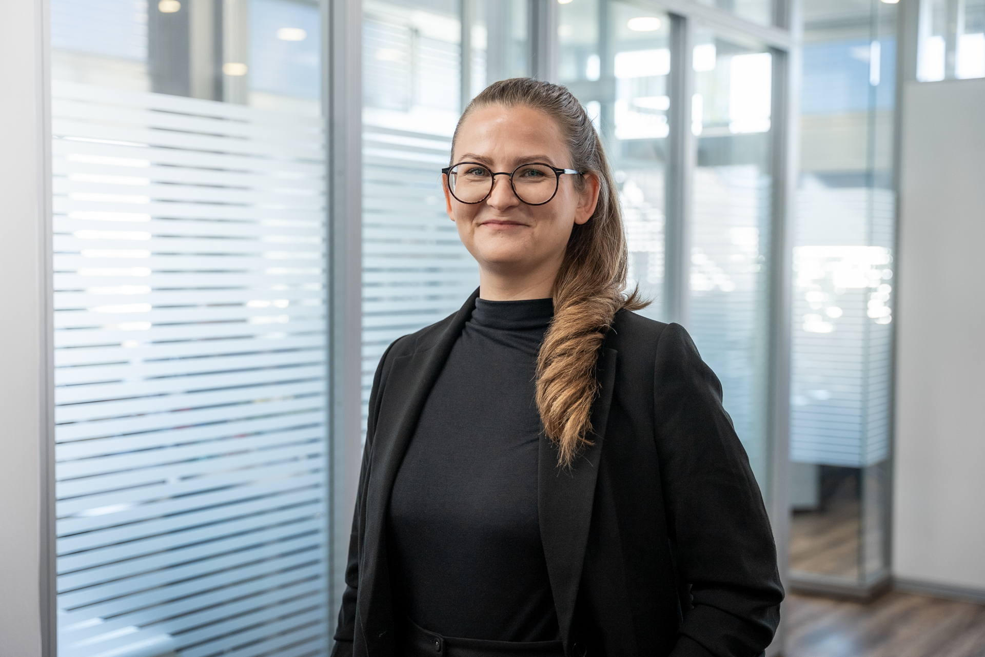 Eine Frau mit langen, gezopfteen Haaren und einer schwarzen Bluse sowie einem Blazer steht in einem modernen Büro mit Glaswänden und Holzboden. Sie lächelt und schaut in die Kamera.