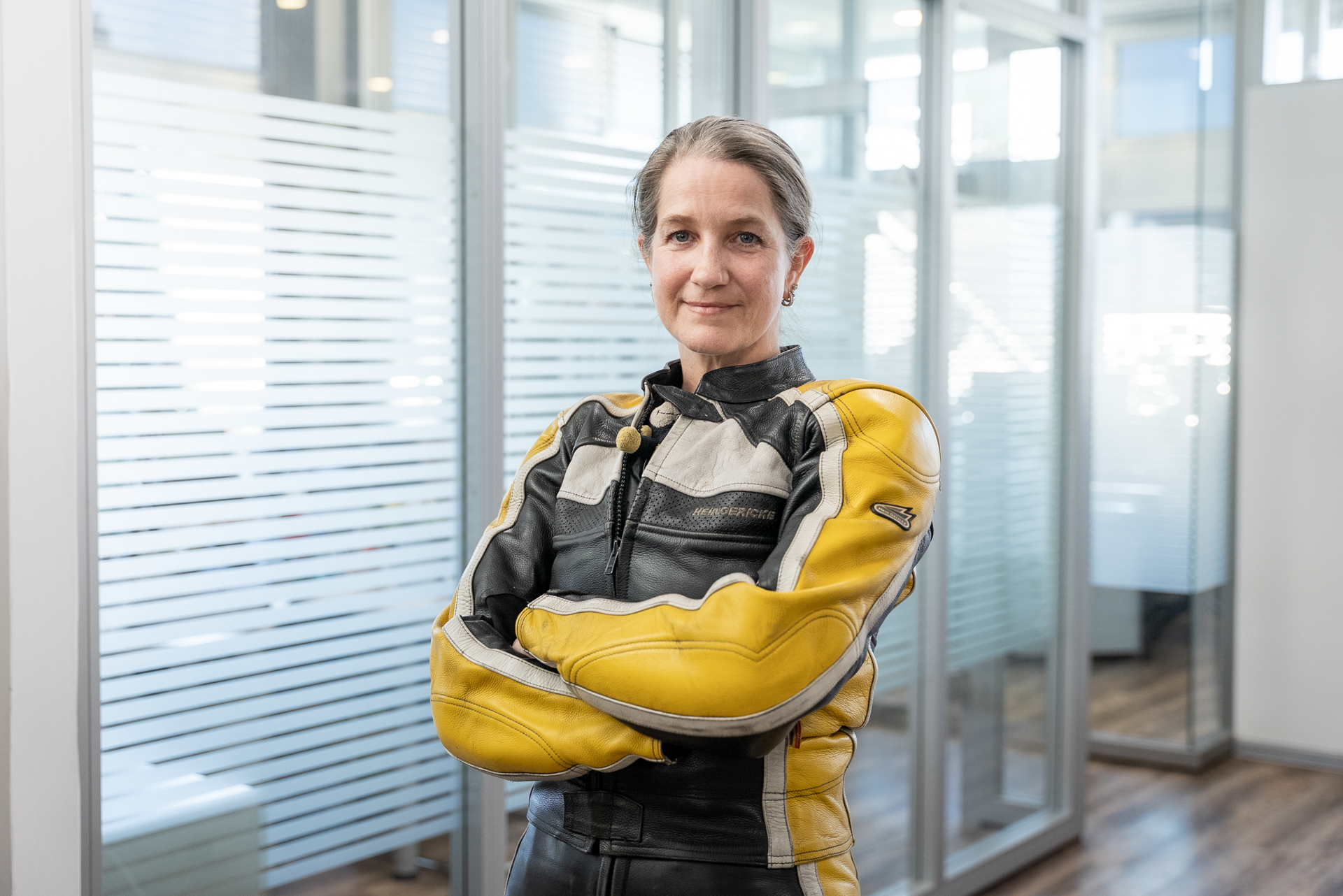 Eine Frau mit kurzen Haaren steht mit verschränkten Armen in einem modernen Büro. Sie trägt eine schwarze und gelbe Motorradjacke und blickt selbstbewusst in die Kamera. Im Hintergrund sind Glaswände sichtbar, die den Raum abtrennen.