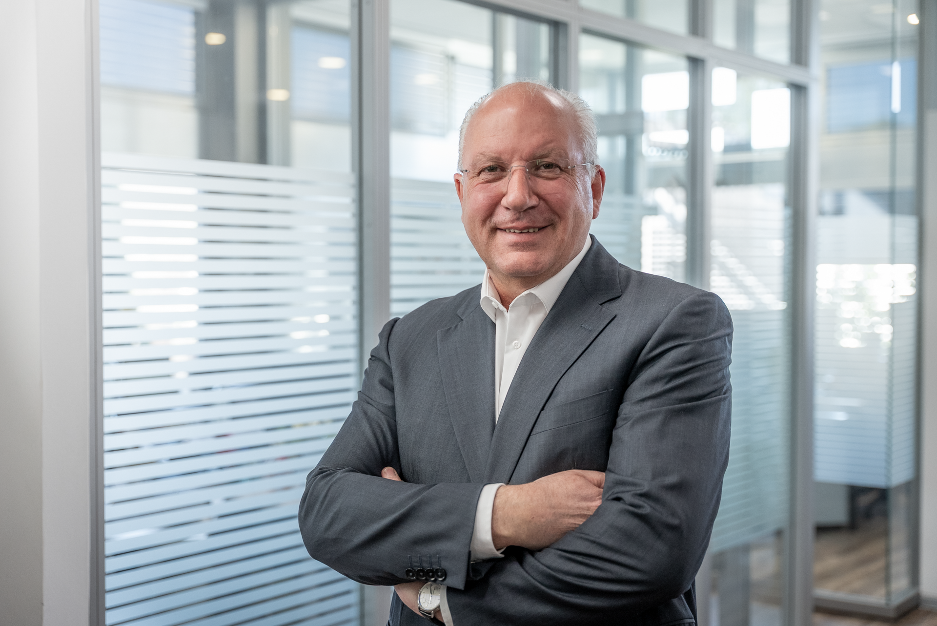 Ein lächelnder Mann in einem grauen Anzug steht mit verschränkten Armen vor einer Glaswand mit durchsichtigen Streifen in einem modernen Büro.