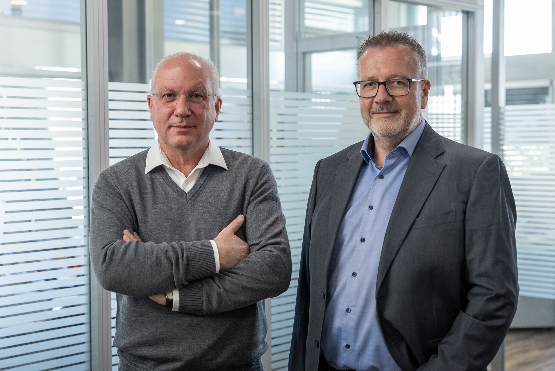 Zwei Männer stehen nebeneinander in einem modernen Büro mit transparenten Fensterscheiben. Der Mann links trägt einen grauen Pullover und hat die Arme verschränkt, während der Mann rechts einen dunklen Anzug und ein hellblaues Hemd trägt. Beide sehen direkt in die Kamera.