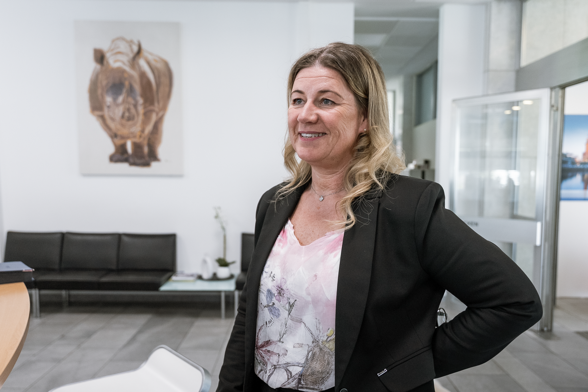 Eine lächelnde Frau im Business-Look steht in einem modernen Empfangsbereich, hinter ihr ist ein Bild eines Nashorns an der Wand zu sehen.