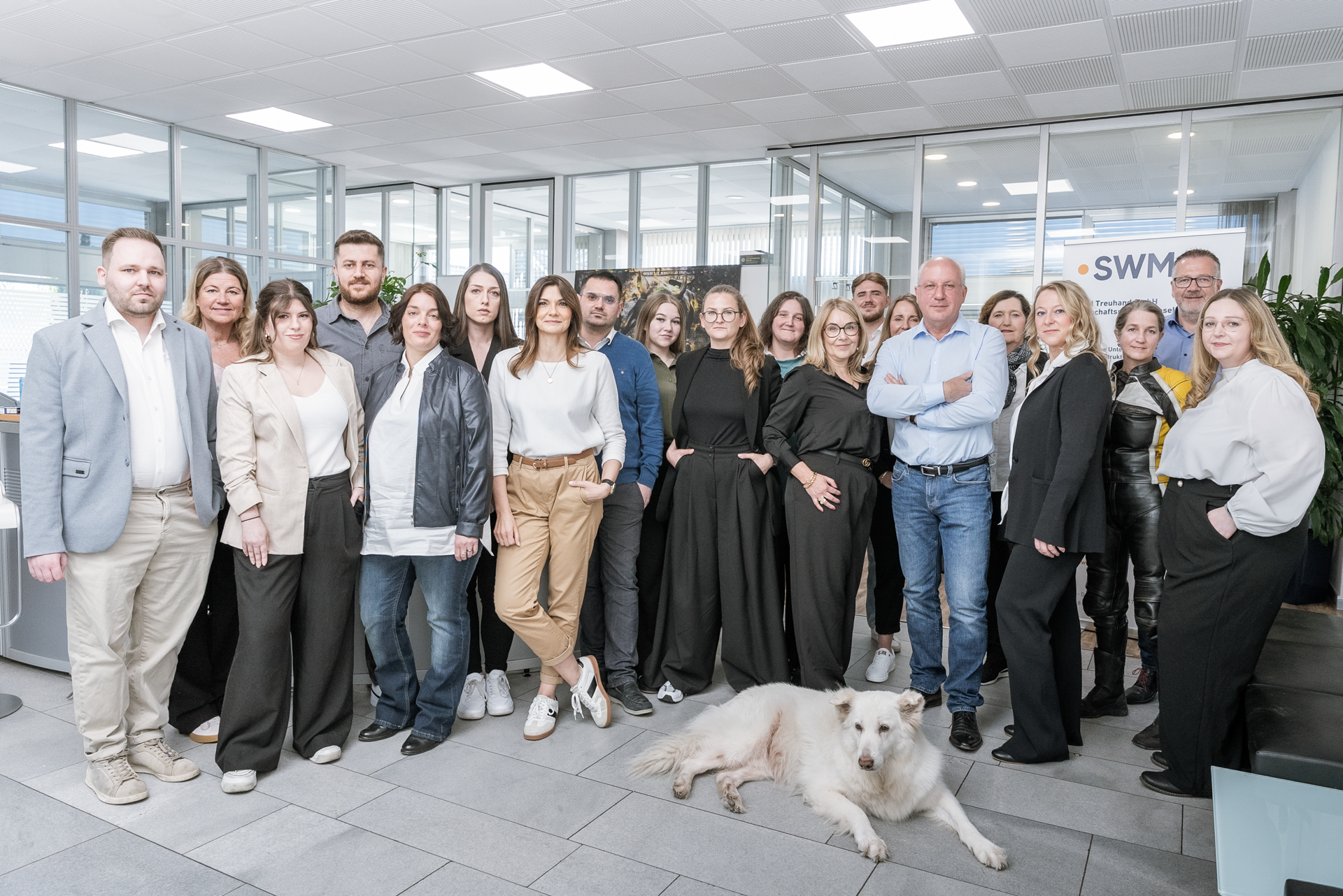 Eine Gruppe von 20 Personen steht in einem modernen Büro mit großen Fenstern, dabei ist ein Hund zu sehen. Die Menschen tragen unterschiedliche professionelle Kleidung und lächeln in die Kamera, einige stehen zusammen, während andere einzeln posieren.