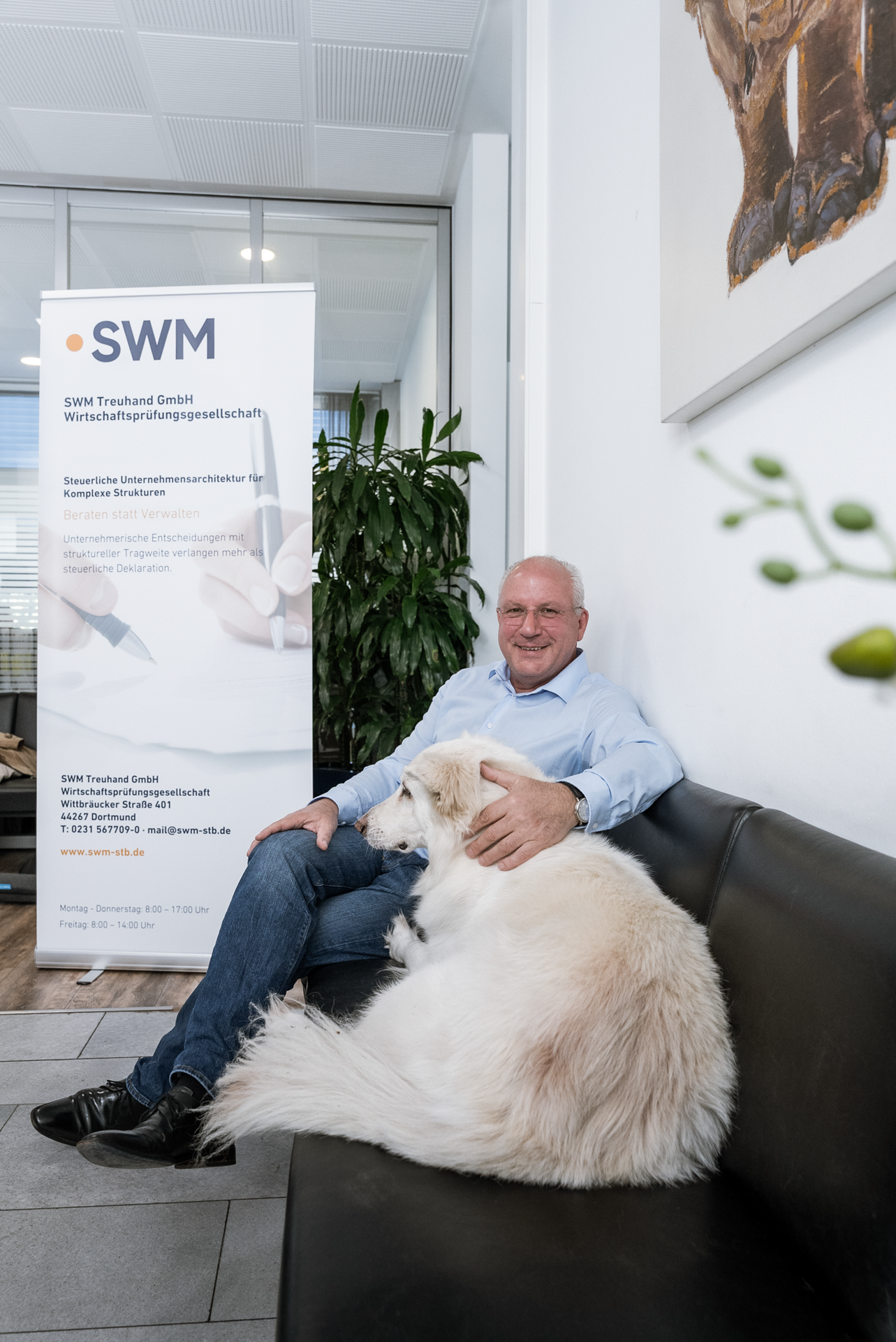 Ein Mann in einem hellblauen Hemd sitzt auf einem schwarzen Sofa in einem modernen Büro, während ein großer, flauschiger Hund entspannt neben ihm sitzt. Im Hintergrund ist ein Banner mit dem Logo „SWM“ und Informationen über die Firma zu sehen.