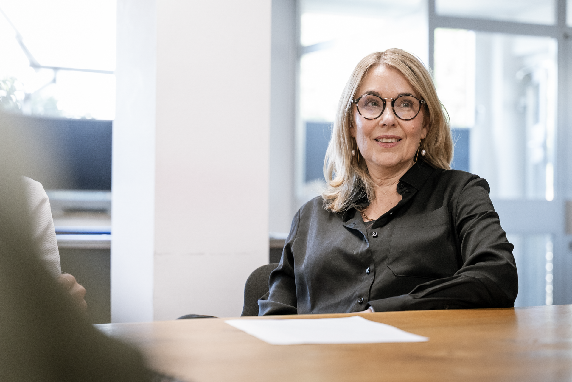 Eine Frau mit Brille sitzt an einem Tisch in einem modern eingerichteten Raum und lächelt, während sie an einem Gespräch teilnimmt. Auf dem Tisch liegt ein weißes Blatt Papier.