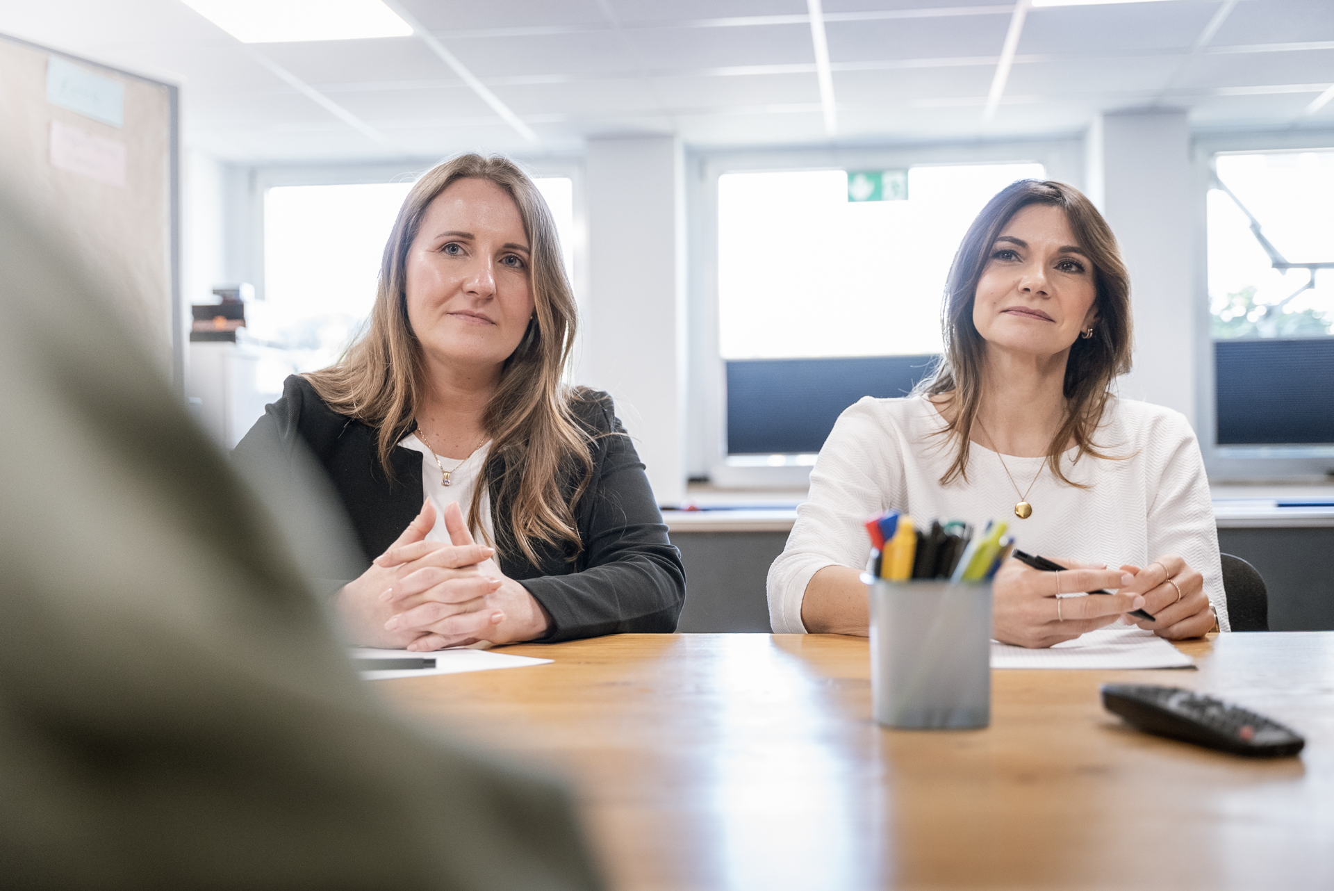 Zwei Frauen sitzen an einem Tisch in einem modernen Büro und hören aufmerksam zu, während jemand außerhalb des Bildes spricht. Vor ihnen steht ein Behälter mit verschiedenen Schreibutensilien.