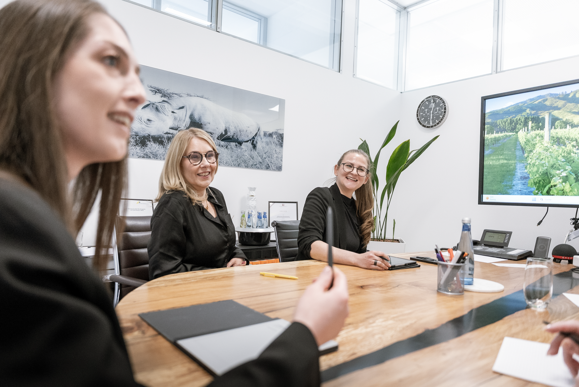 Drei Frauen in formeller Kleidung sitzen an einem Konferenztisch in einem modernen Besprechungsraum und diskutieren. Im Hintergrund ist ein Bild einer Landschaft sowie eine Uhr sichtbar. Auf dem Tisch stehen Schreibutensilien und Gläser mit Wasser.
