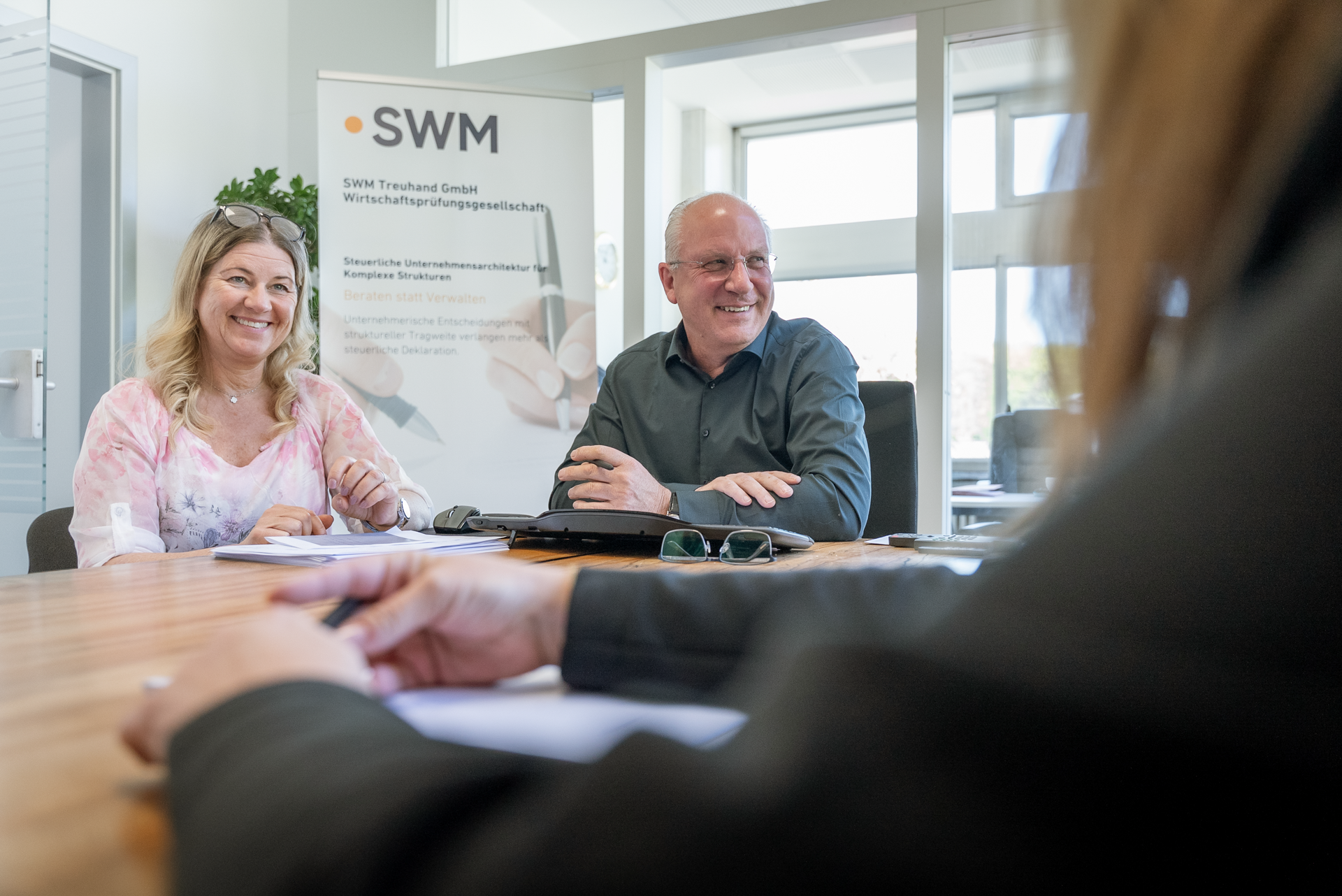 Zwei Personen sitzen an einem Tisch in einem Büro, lächeln und führen ein Gespräch, während eine dritte Person außerhalb des Bildes anscheinend zuhört. Im Hintergrund ist ein Plakat mit der Aufschrift "SWM" und weiteren Informationen sichtbar.