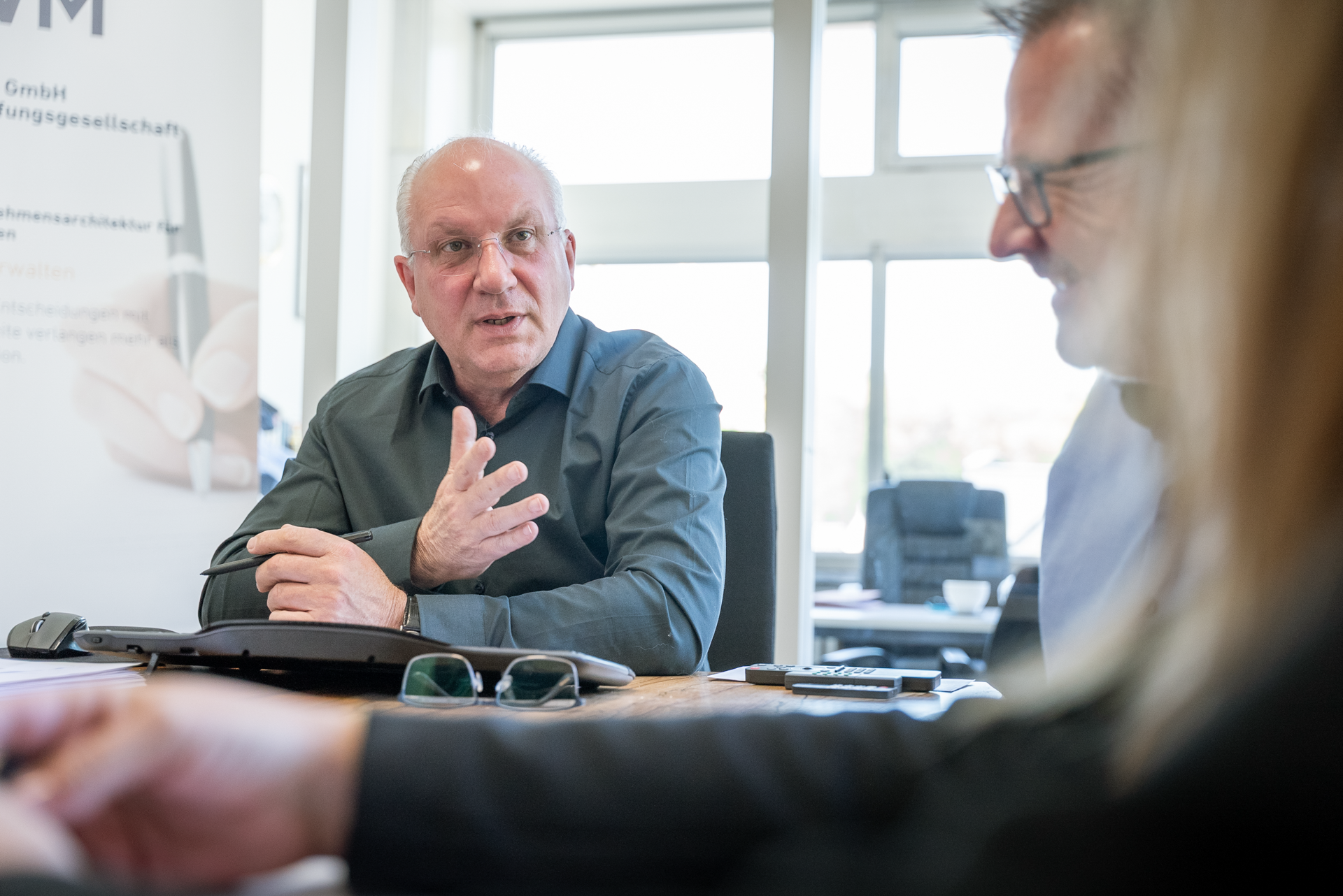 Ein älterer Mann mit Brille und einem grauen Hemd sitzt am Tisch und gestikuliert, während er spricht. Auf der linken Seite sind Teile eines weiteren Gesprächsteilnehmers und einige Büromaterialien sichtbar. Im Hintergrund sind Büromöbel und Tageslicht durch große Fenster zu sehen.