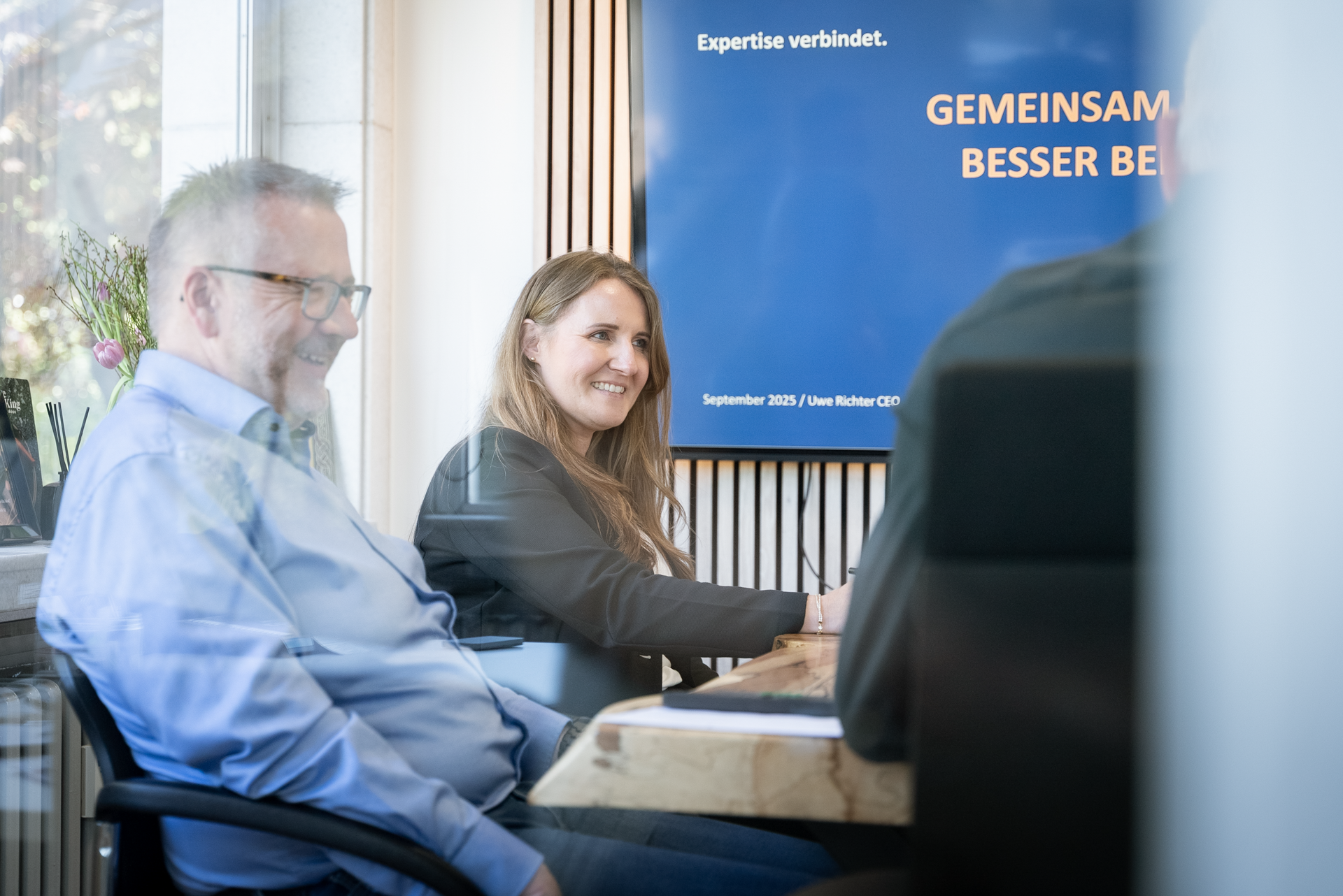 In einem Besprechungsraum sitzen ein Mann und eine Frau an einem Tisch und diskutieren. Die Frau lächelt, während der Mann einen freundlichen Gesichtsausdruck hat. Im Hintergrund ist ein Banner mit dem Text „Expertise verbindet“ und „GEMEINSAM BESSER BEI“ zu sehen.