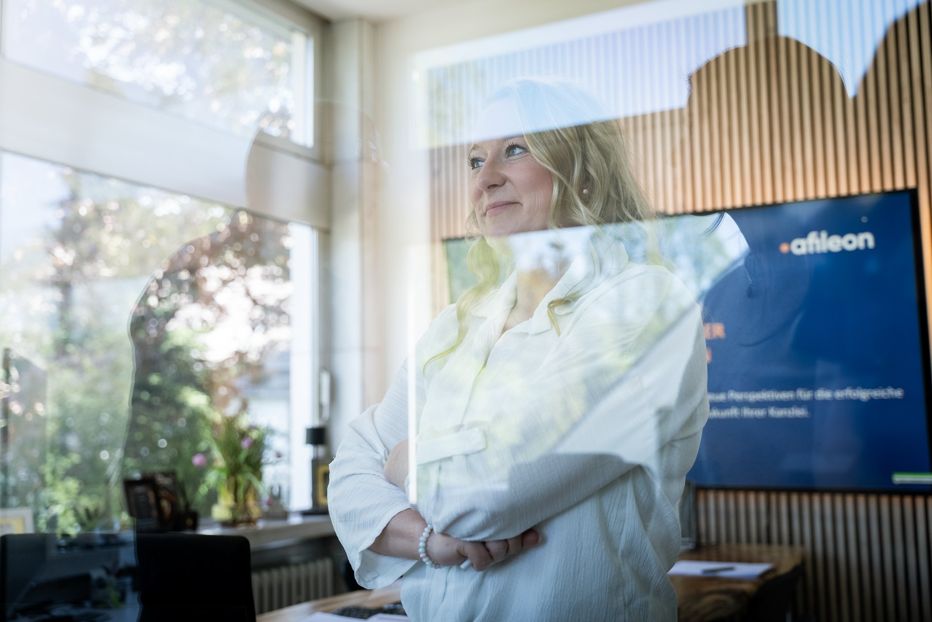 Eine Person mit blonden, lockigen Haaren steht mit verschränkten Armen in einem modernen, hellen Raum mit großen Fenstern, hinter denen man Bäume sehen kann. Im Hintergrund ist ein Bildschirm zu sehen, auf dem Informationen über ein Unternehmen präsentiert werden.