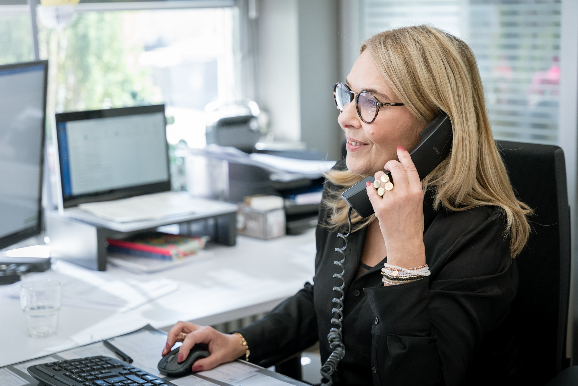 Eine Frau mit blonden Haaren und Brille sitzt an einem Schreibtisch und spricht am Telefon, während sie auf einen Computerbildschirm schaut. In ihrer Nähe sind Unterlagen und ein Glas Wasser sichtbar.