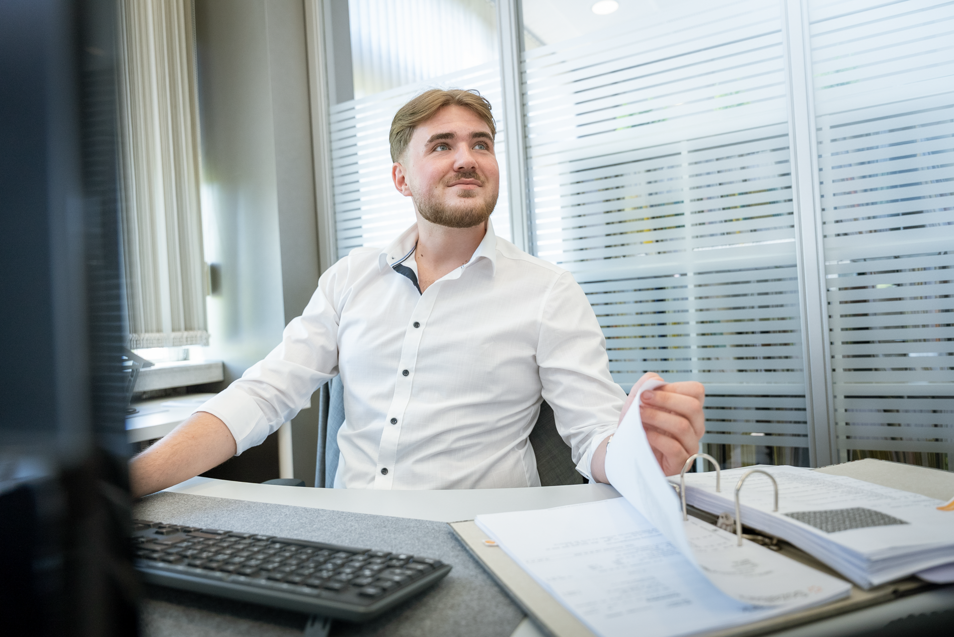 Ein Mann mit blonden Haaren sitzt an einem Schreibtisch, trägt ein weißes Hemd und schaut nachdenklich in die Ferne. Auf dem Tisch liegen mehrere Dokumente und ein Computer ist sichtbar.