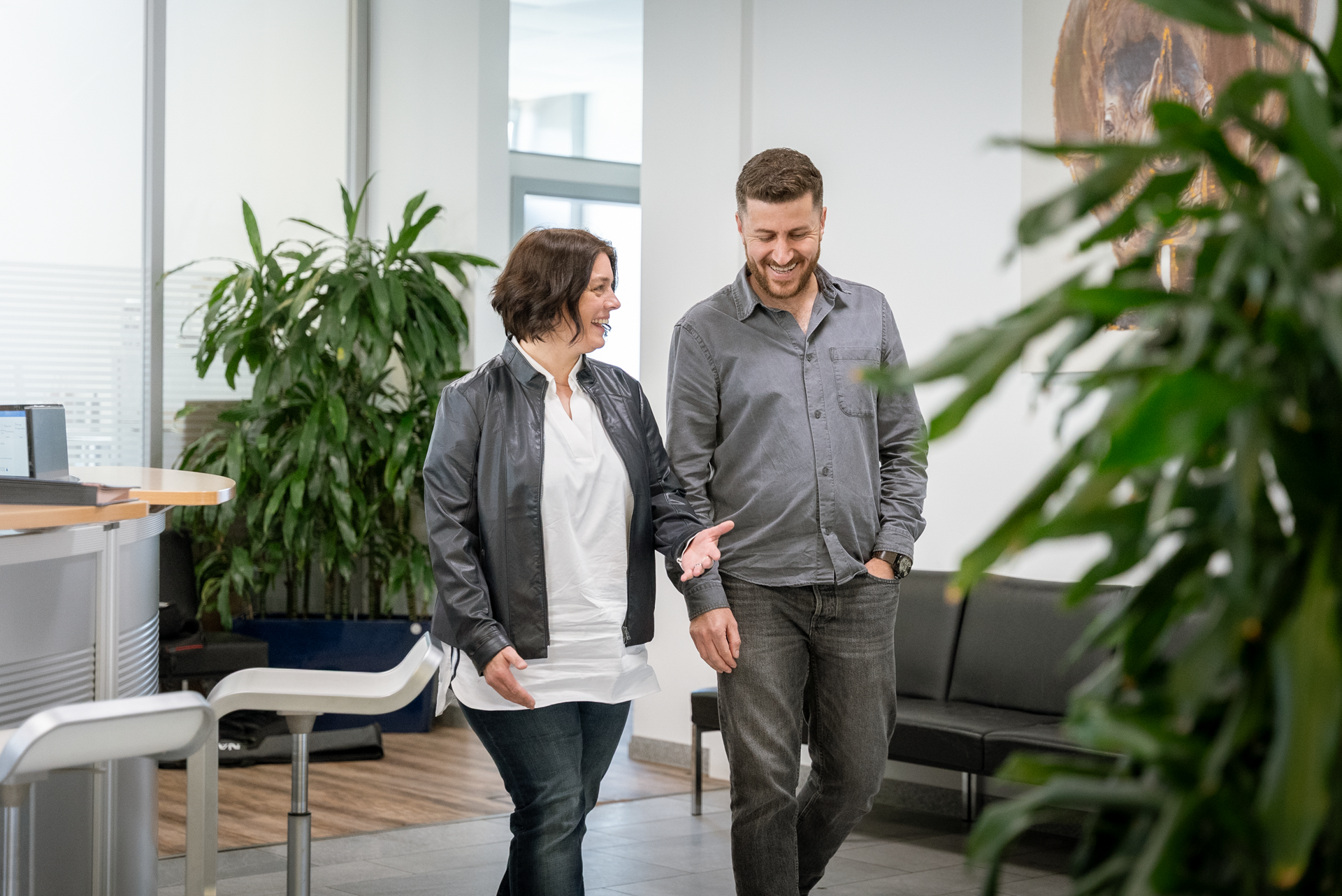 Ein Mann und eine Frau in lässiger Kleidung gehen in einem hellen, modernen Bürogebäude aufeinander zu und unterhalten sich, während sie an einer großen Pflanze vorbeigehen.