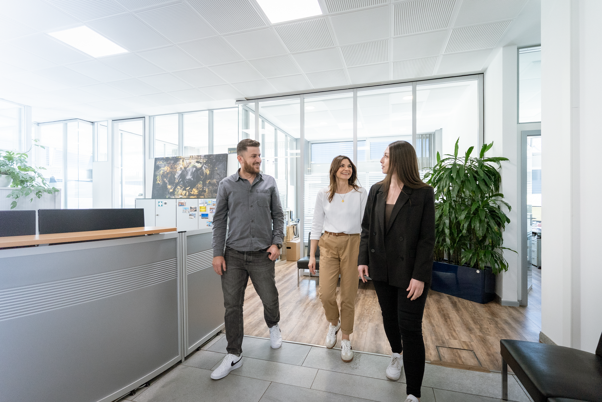 Drei Menschen, zwei Frauen und ein Mann, gehen gemeinsam in einem modernen Büro mit großen Fenstern und Pflanzen. Die Frauen tragen lässige Kleidung, während der Mann ein Hemd und eine Hose trägt.