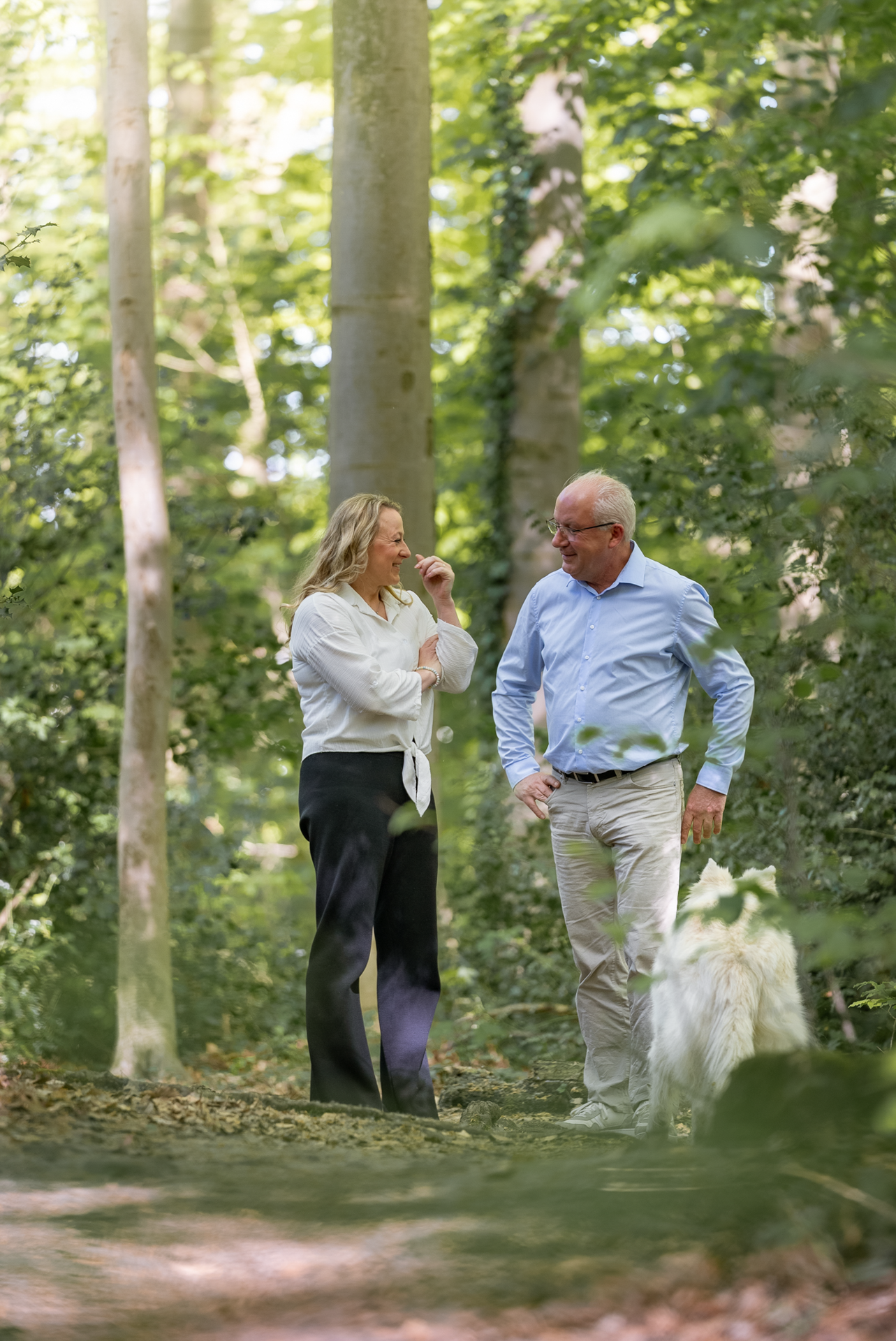 Ein Mann und eine Frau unterhalten sich im Wald, umgeben von grünen Bäumen. Die Frau trägt eine weiße Bluse und schwarze Hose, während der Mann ein hellblaues Hemd und beige Hosen anhat. Ein Hund ist ebenfalls im Bild sichtbar.