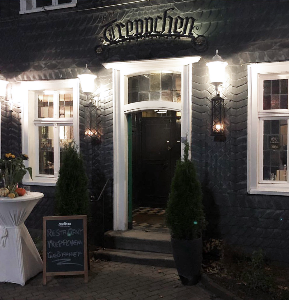 Der Eingang eines Restaurants mit dem Namen "Clemenschen" ist sichtbar, umgeben von eleganten, beleuchteten Wandleuchten. Vor der Tür stehen zwei kleine Pflanzen und ein Schild mit der Aufschrift "Restaurant", daneben ein Tisch mit Blumenarrangement.